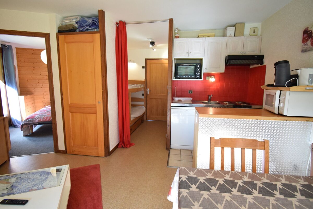 A compact apartment interior featuring a kitchenette with red tiles, a dining area, and glimpses of a bedroom and bunk beds, suggesting a practical, small living space.