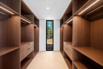 Sleek walk-in closet with dark wood shelving, drawers, and accent lighting. A narrow window at the far end reveals outdoor greenery.