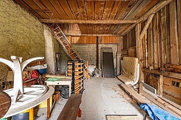 A rustic, cluttered storage room or attic with exposed wooden beams, a ladder leading up, and various discarded furniture and items.