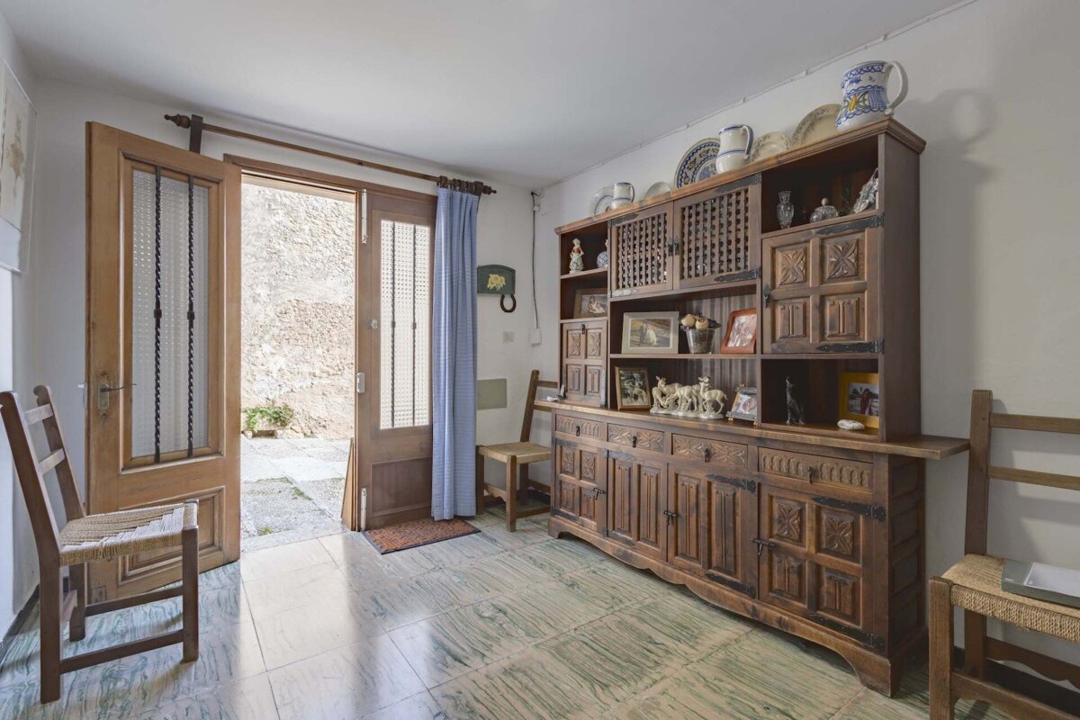 A rustic room with an open wooden door leading to a courtyard. A large, ornate wooden hutch with decor sits on a greenish tiled floor, with chairs nearby.