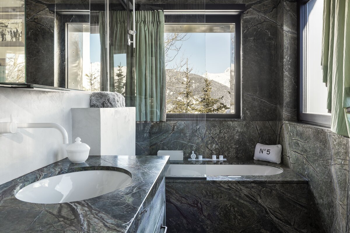 Modern dark marble bathroom with a sink, bathtub, and large window revealing a snowy mountain and pine tree landscape.