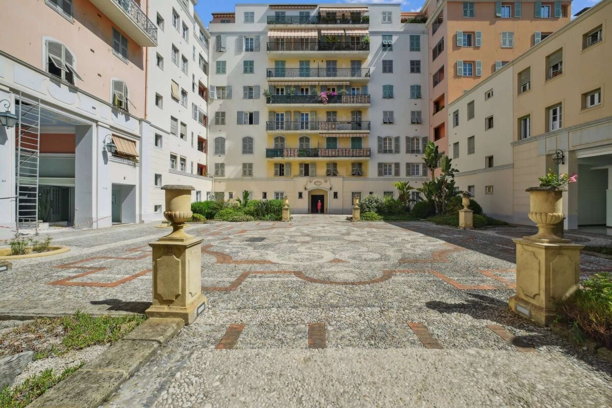A residential courtyard featuring an elaborate pebble mosaic ground, flanked by colorful multi-story buildings with balconies, shutters, and decorative urns.
