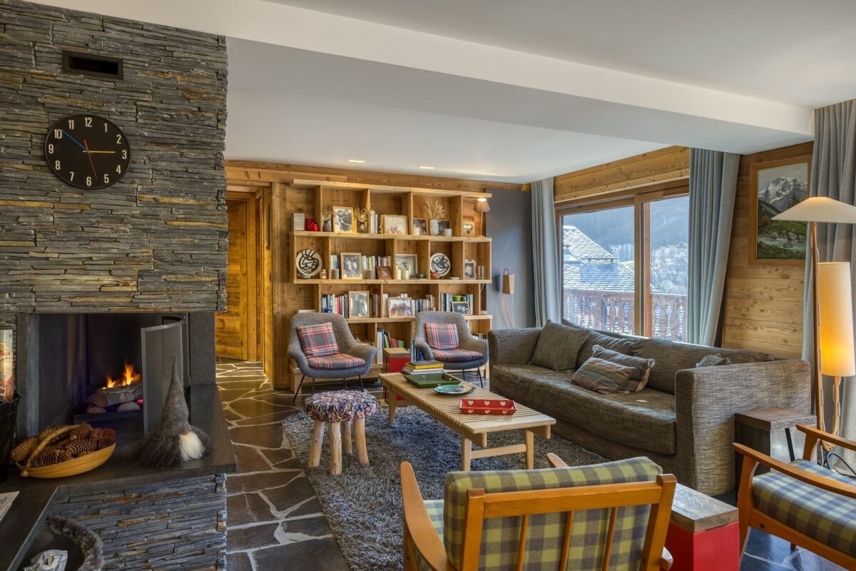 A cozy, rustic living room featuring a stone fireplace, wooden walls, ample seating, and a large window overlooking a scenic mountain or village view.