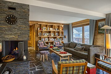 A cozy, rustic living room featuring a stone fireplace, wooden walls, ample seating, and a large window overlooking a scenic mountain or village view.
