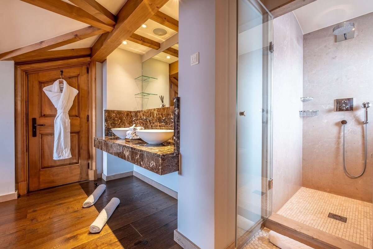 A well-appointed bathroom features a marble double vanity, a glass walk-in shower, rich wooden floors, exposed beams, and a bathrobe.