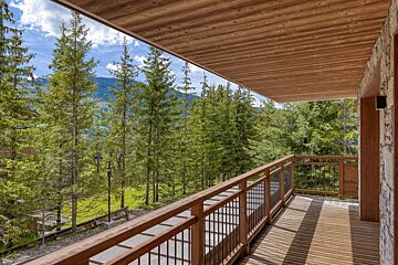 A wooden balcony offers a beautiful view of a dense pine forest and distant mountains under a bright, partly cloudy sky.