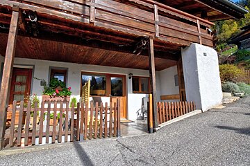 Rustic chalet with a wooden balcony above a fenced patio with flowers, on a hillside beside an asphalt road.