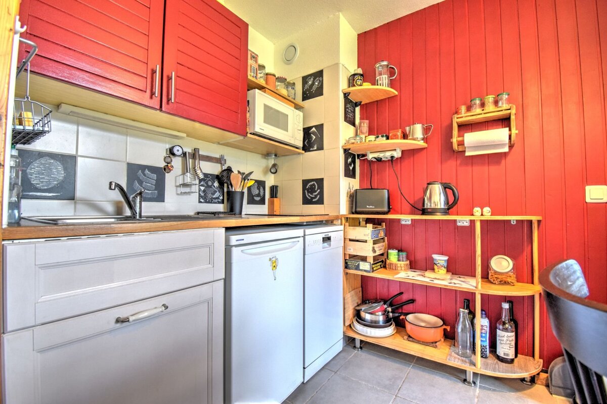 A compact kitchen features striking red paneled walls & upper cabinets contrasting with light gray lower cabinets, full of small appliances & wooden shelves.