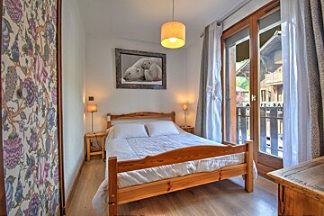 A cozy, rustic bedroom featuring a wooden bed, a polar bear painting, floral wallpaper, and large glass doors opening to a balcony.