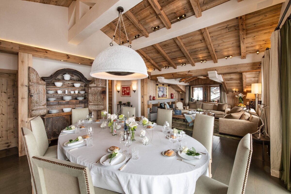 A luxurious chalet dining room with an oval table set for a meal, a large dome pendant light, and exposed wood ceilings.