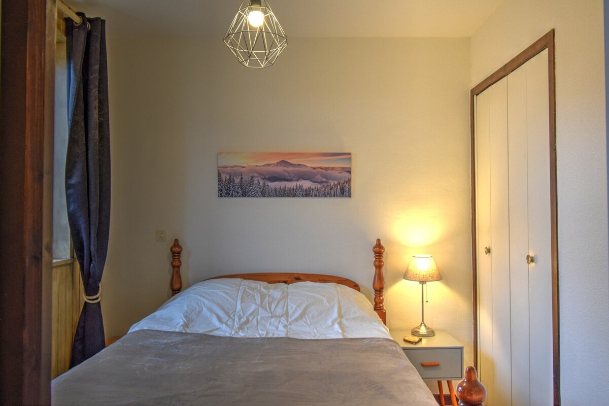Cozy bedroom with a wooden bed, mountain artwork above, a lit bedside lamp on a nightstand, dark window curtains, and a white wardrobe.