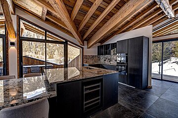 Luxury kitchen with dark cabinets, granite countertops, exposed wooden beams, and expansive windows showcasing a snowy mountain and forest view.