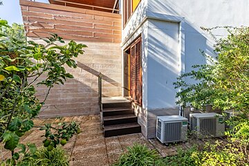 A building's exterior features brown stairs to a shuttered door, stone and white walls, lush green foliage, and two outdoor AC units.