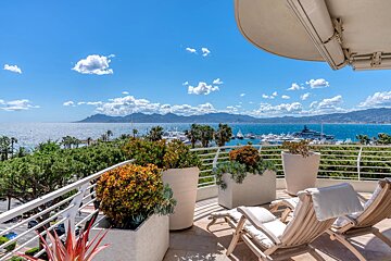 Sunny balcony overlooking a sparkling blue sea with yachts and distant mountains, under a bright sky, featuring lounge chairs and lush potted plants.