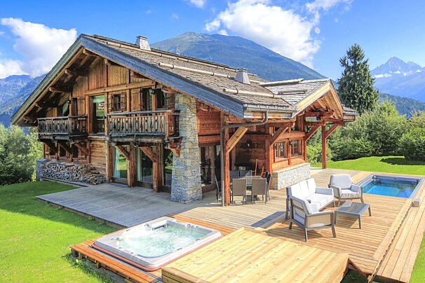 A luxurious wooden chalet with a large deck, outdoor furniture, hot tub, and pool, nestled amidst green grass and majestic mountains under a blue sky.
