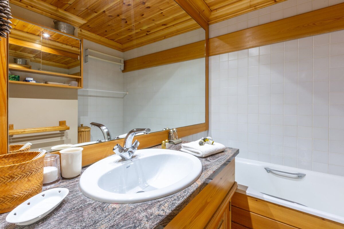 Cozy rustic bathroom with a wooden ceiling, white tiled walls, granite counter with a sink, large mirror, and a bathtub.