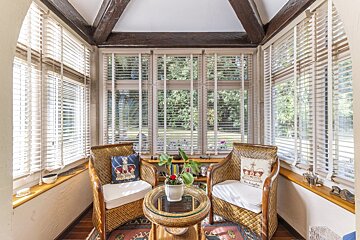 A cozy sunroom with wicker chairs & a table, large windows with blinds, exposed beams, & a garden view.