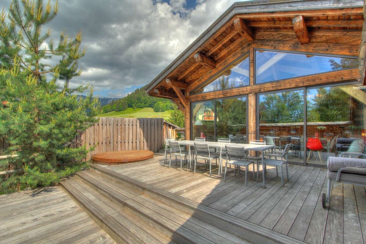 A modern timber-frame chalet with large glass walls, a spacious wooden deck with dining area and hot tub, overlooking a cloudy mountain view.