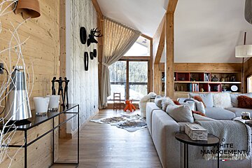 A spacious, modern chalet living room with rustic wood and beams, a plush gray sofa, and a large window offering scenic snowy mountain views.