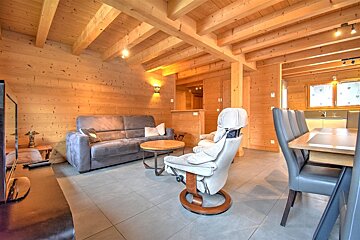 A cozy, modern cabin interior with warm wood walls, ceiling beams, grey sofa, recliner, dining table with chairs, and tiled floors.