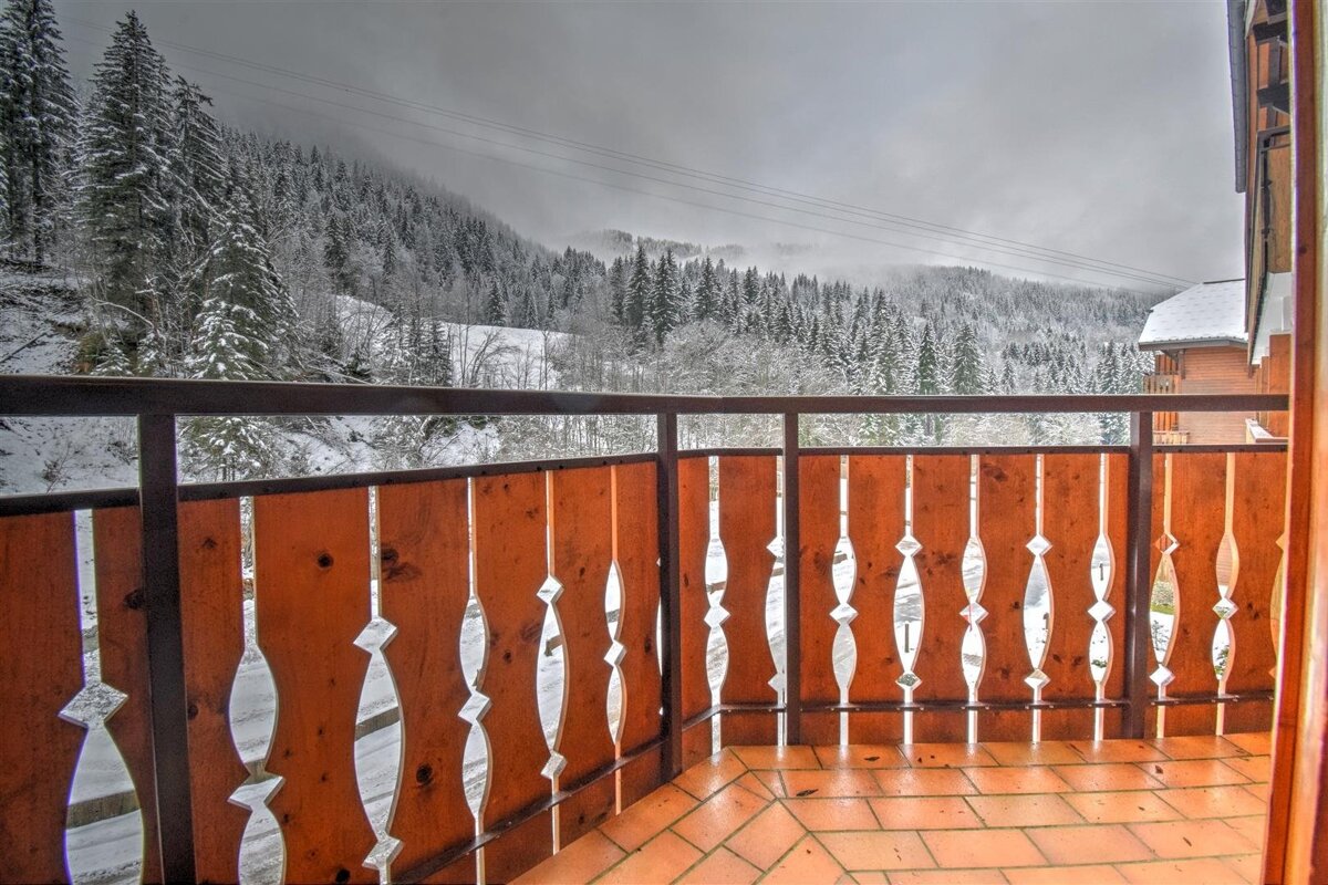 View from a tiled balcony with a wooden railing looking out at a snowy pine forest under a grey winter sky.