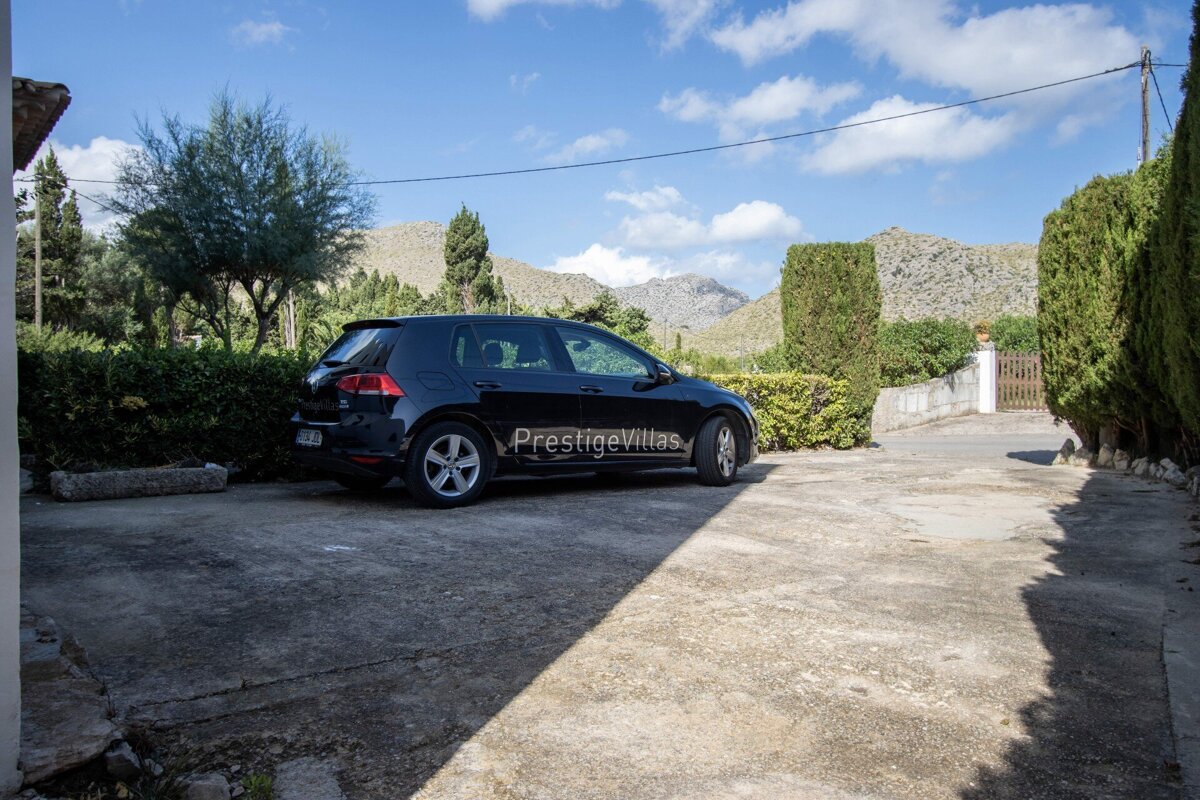 A black car is parked on a concrete driveway, surrounded by green hedges and trees, with mountains and a blue sky in the background.