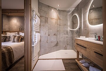 Modern bathroom with stone tiles, a glass shower, and a wooden vanity next to a cozy chalet bedroom.
