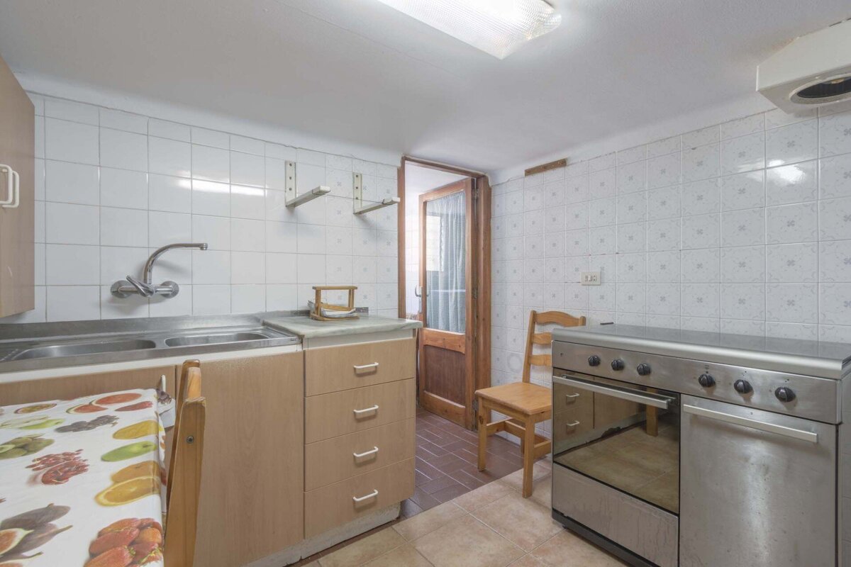 A dated kitchen with white tiled walls, a double sink, wooden cabinets, a stainless steel stove, and a wooden chair near an open door.