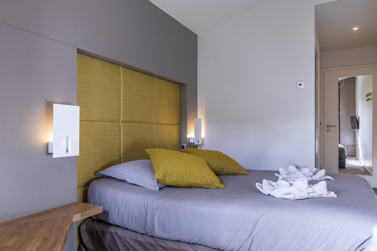 A modern bedroom with a grey-sheeted bed, yellow pillows, a matching yellow headboard, and sleek wall sconces.