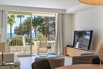 A modern living room with a balcony overlooking the sea and palm trees. White furnishings, a glass table, and a large TV complete the bright space.