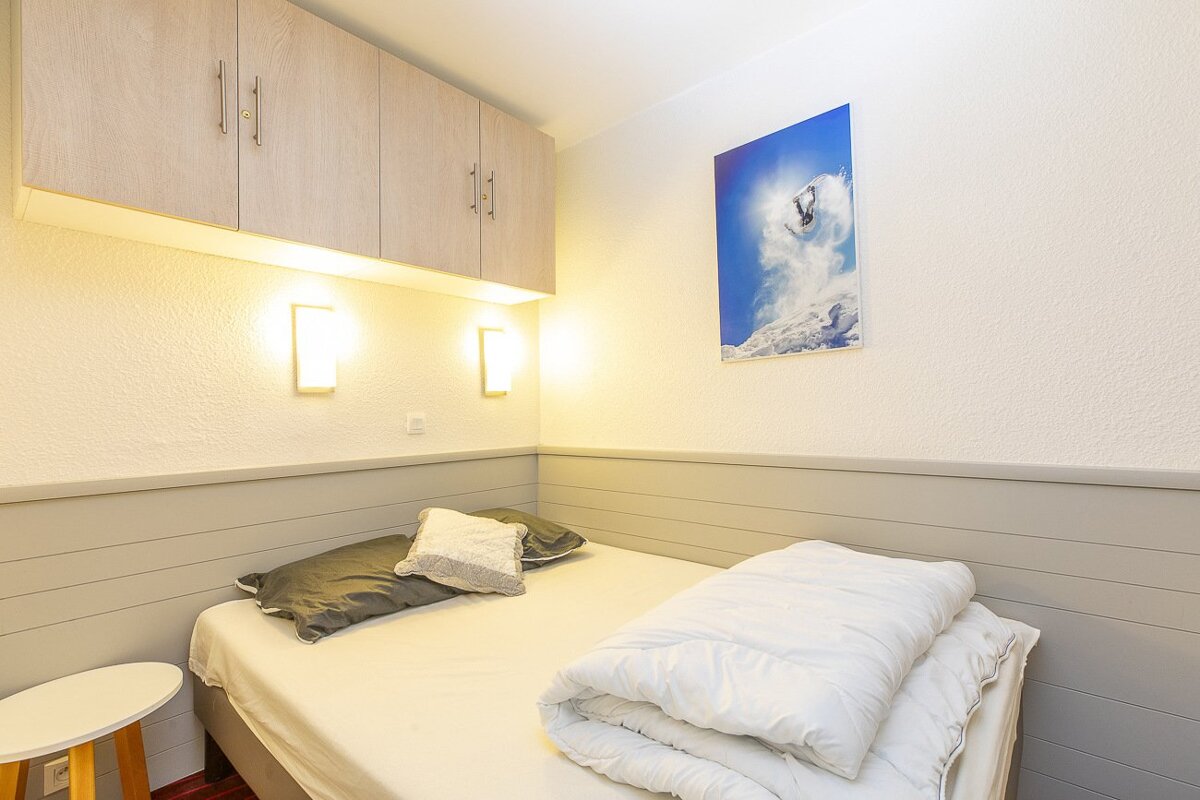 A compact bedroom features a bed with white bedding, light wood overhead cabinets, wall sconces, and a snowboarder print. Walls are white and grey paneled.