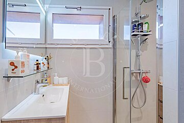 A modern, well-lit bathroom featuring a white sink, mirror, toiletries on shelves, and a glass-enclosed shower with a window above the vanity.