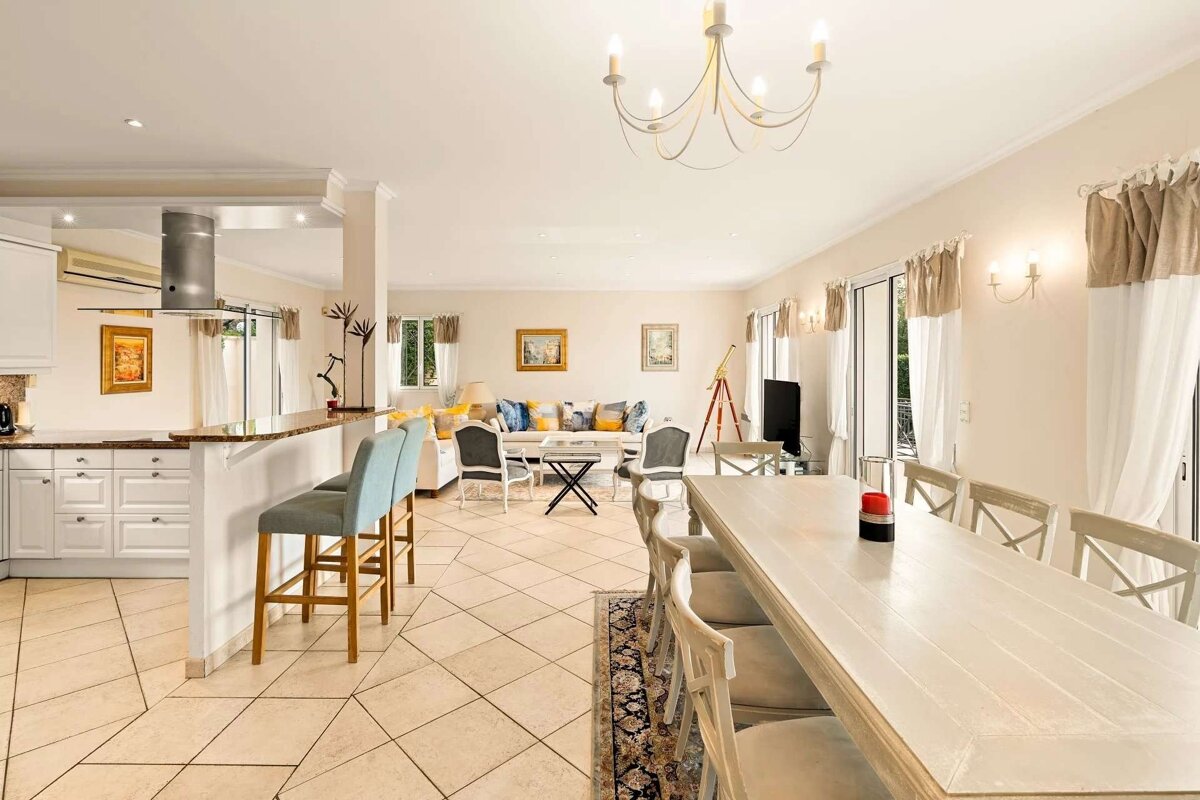 A bright open-plan kitchen, living, and dining room with light decor, tiled floor, and bar seating.