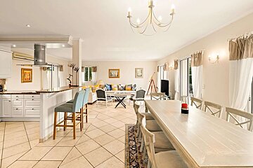 A bright open-plan kitchen, living, and dining room with light decor, tiled floor, and bar seating.