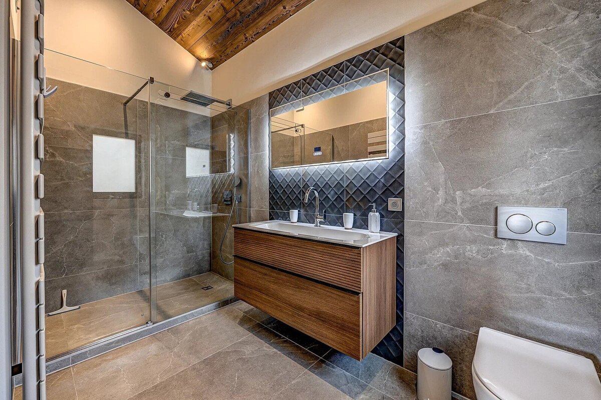 Modern bathroom featuring grey stone-look tiles, a glass shower cabin, a floating wooden vanity, and a rustic wood ceiling.