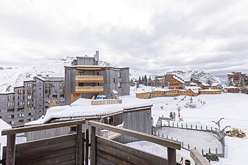 Snowy alpine village with timber buildings, an outdoor ice rink, and cloudy mountains. A snowplow is near the rink.