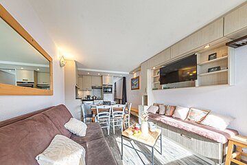 Bright, open-plan living space featuring a brown sofa, coffee table, dining area, compact kitchen, and a built-in TV unit with shelving.