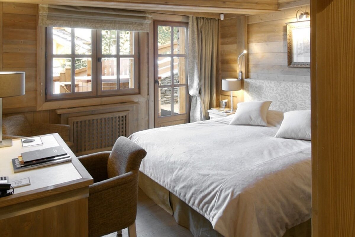 A warm, wood-paneled bedroom featuring a large bed with white linens, a desk, and windows/door to an outdoor area, creating a cozy chalet feel.