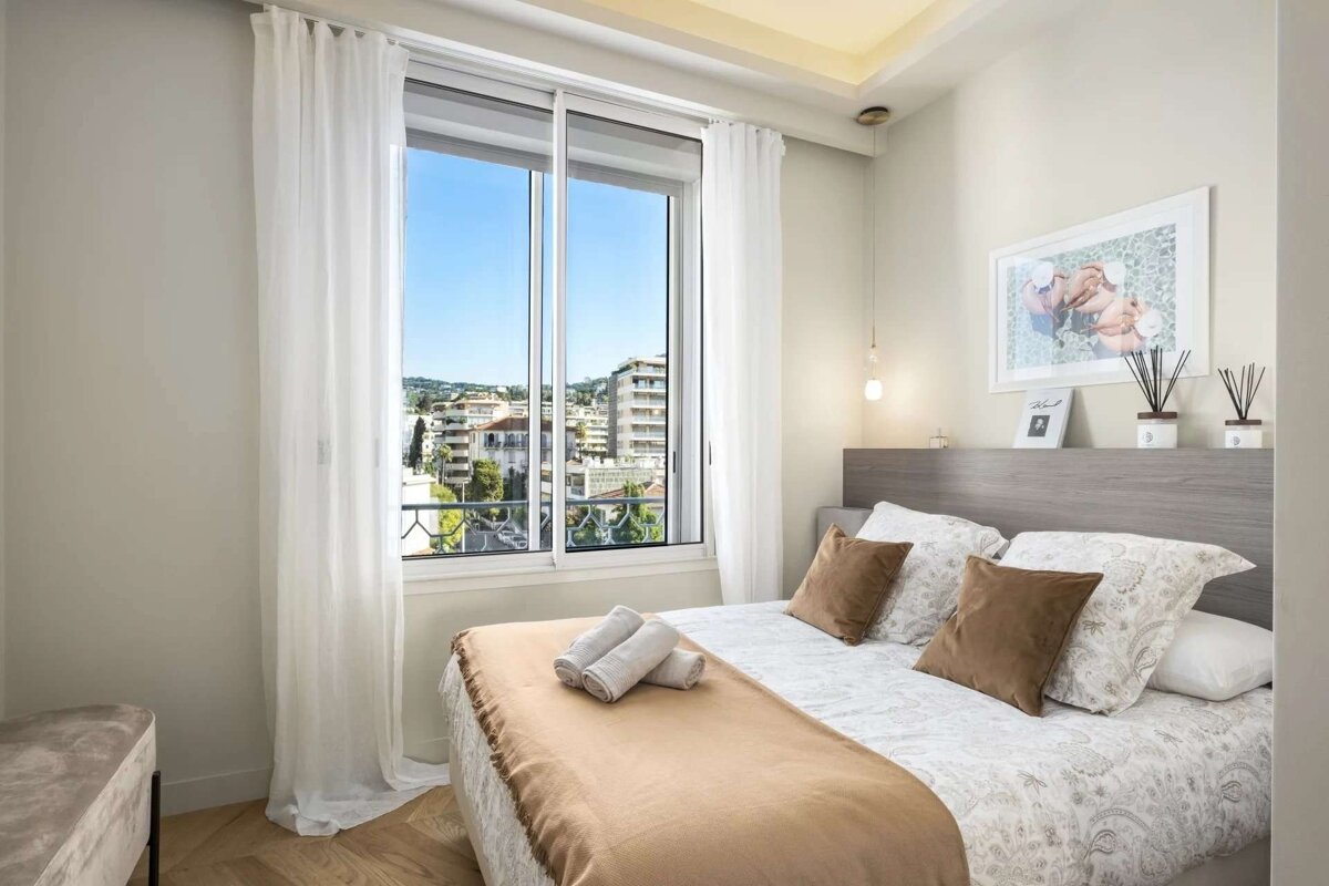 A modern bedroom with a neatly made bed, brown pillows, and a large window offering a vibrant city view, framed by white sheer curtains.