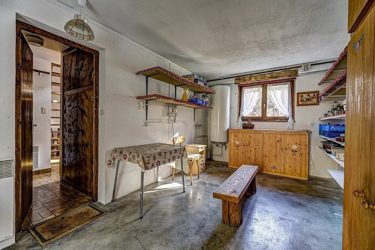 A rustic room with a concrete floor, wooden door leading to stairs, table, bench, shelves, and a window with white curtains.