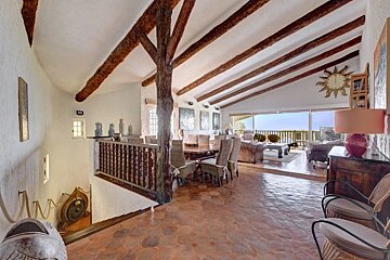 Rustic open-plan interior with exposed ceiling logs, tiled floor, dining set, and living area overlooking a balcony.