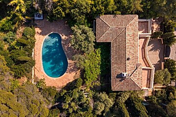 Aerial view of a house with a terracotta tiled roof, a large blue swimming pool, and lush green trees and foliage.
