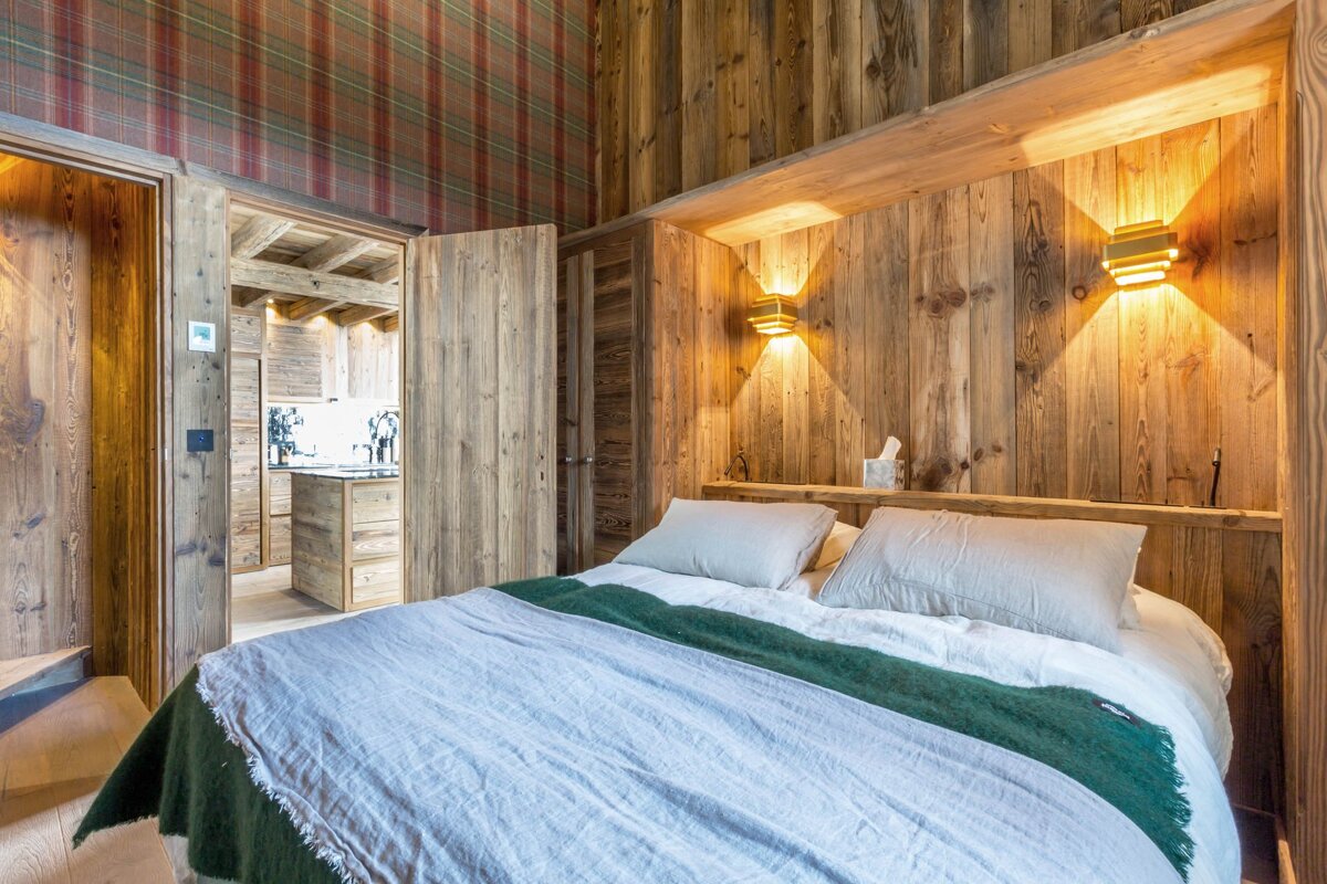 Cozy chalet bedroom featuring rustic wood walls, tartan wallpaper, warm sconces, and a bed with a green throw.