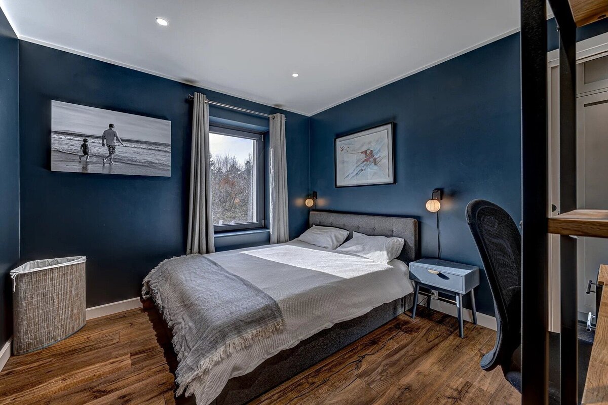 A modern bedroom with deep blue walls, a grey bed, warm wooden floors, a bright window, and framed art, including a beach scene.