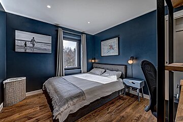 A cozy bedroom with deep blue walls, a gray bed, and warm hardwood floors. Natural light streams from the window, highlighting a black-and-white print.