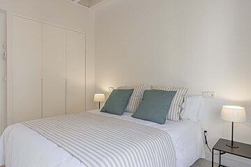 Clean, minimalist bedroom with a bed, striped bedding, teal pillows, two lamps on nightstands, and built-in white wardrobes.