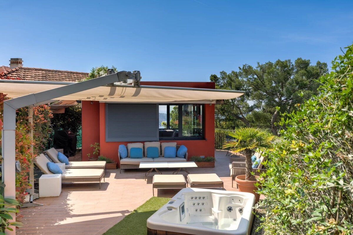 A sunny outdoor patio featuring a hot tub, lounge chairs, and a shaded seating area with blue pillows.