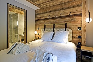 Cozy chalet bedroom with wood paneling, a white double bed, and an adjacent modern bathroom visible through a doorway.