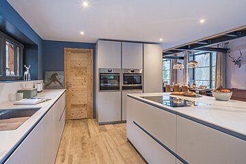 A contemporary kitchen features light grey cabinets, a large island with a hob, wooden flooring, and a blue accent wall. It opens to a dining area with scenic windows.