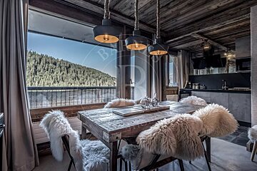 Rustic dining/kitchen with furry chairs, industrial lights, wooden ceiling, and a large window overlooking a forest and lake.